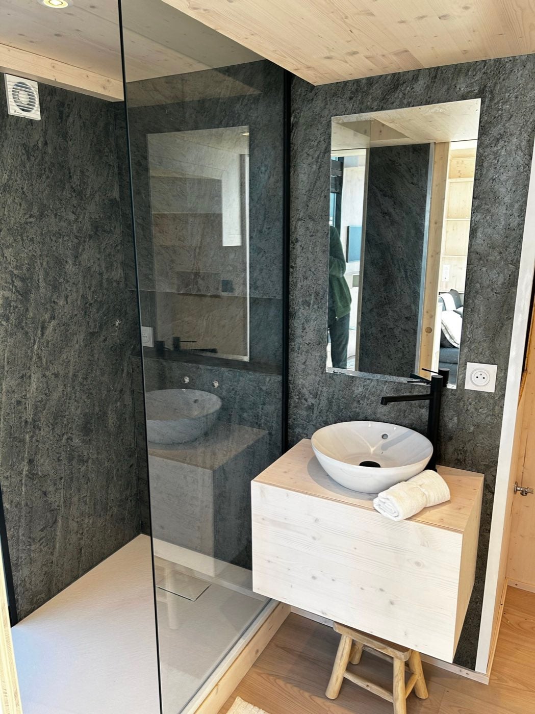 Salle d'eau doublée de parement pierre : douche de plain-pied à paroi vitrée et vasque blanche sur meuble suspendu en épicéa