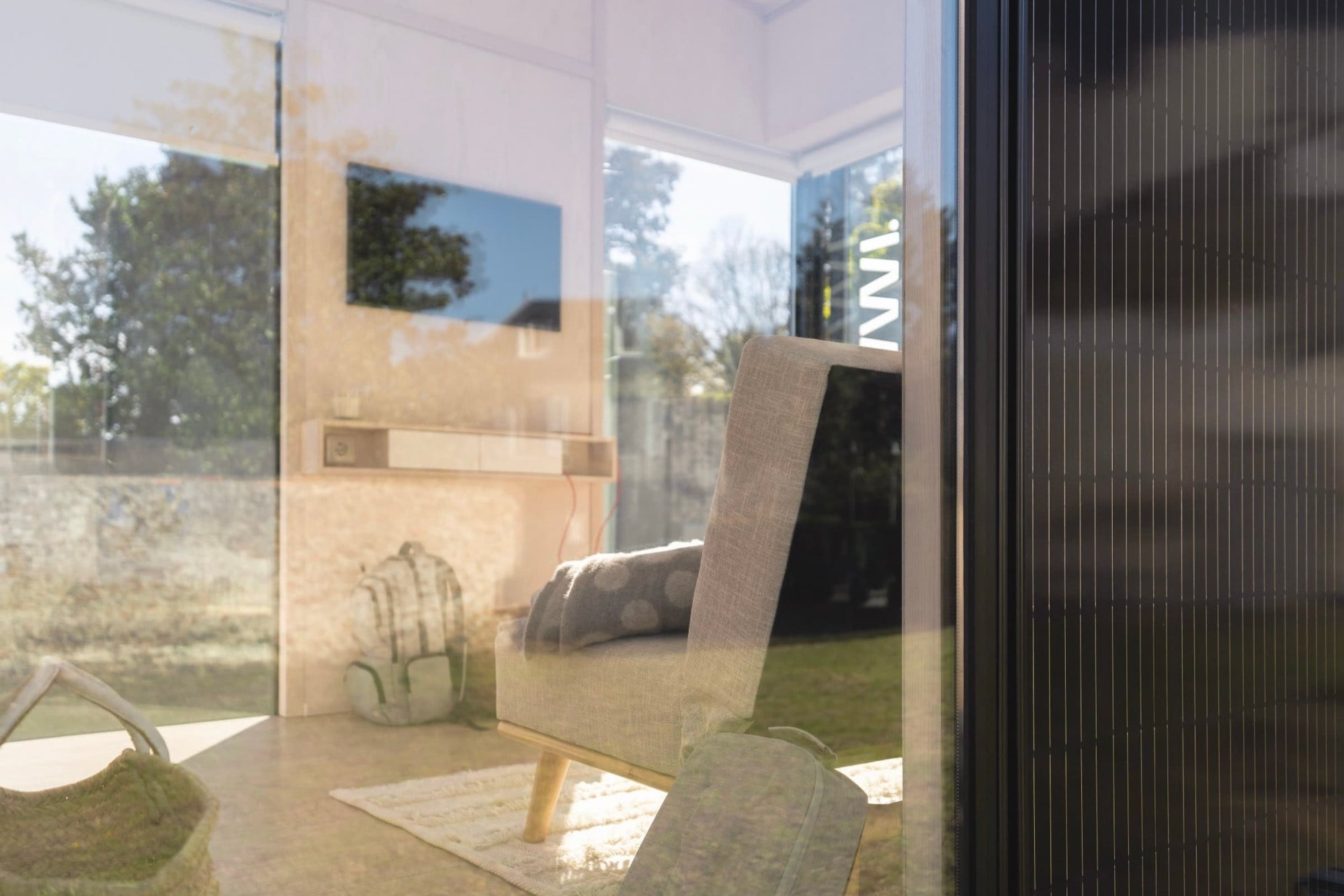 Séjour d'un module Hoummi vu à travers la baie vitrée : fauteuil et plaid, panier et sac à dos au sol, le verre superposant l'intérieur et le reflet du jardin