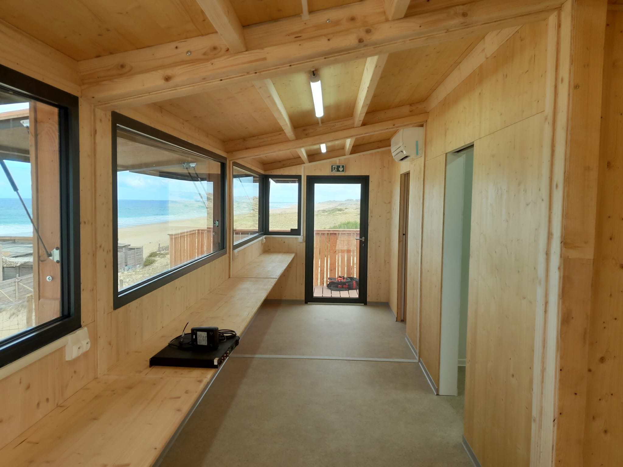 Salle de veille aménagée d'un poste de secours Hoummi : parois et plafond en bois clair, tablette continue et baie filante cadrant la plage