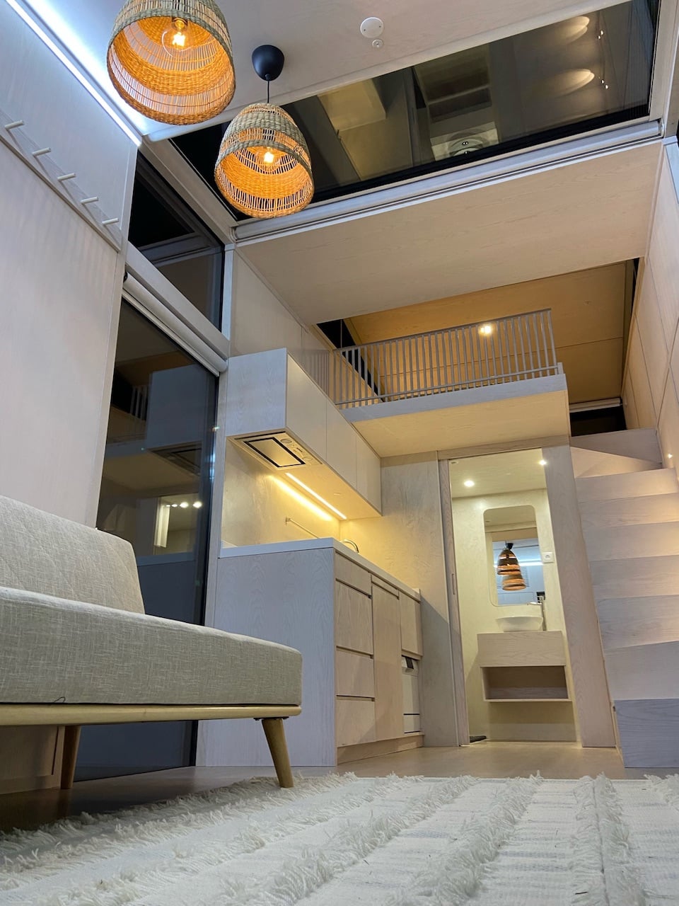 Intérieur d'un studio Hoummi meublé : canapé au premier plan, cuisine équipée, salle d'eau au fond, escalier et chambre en mezzanine sous un toit vitré
